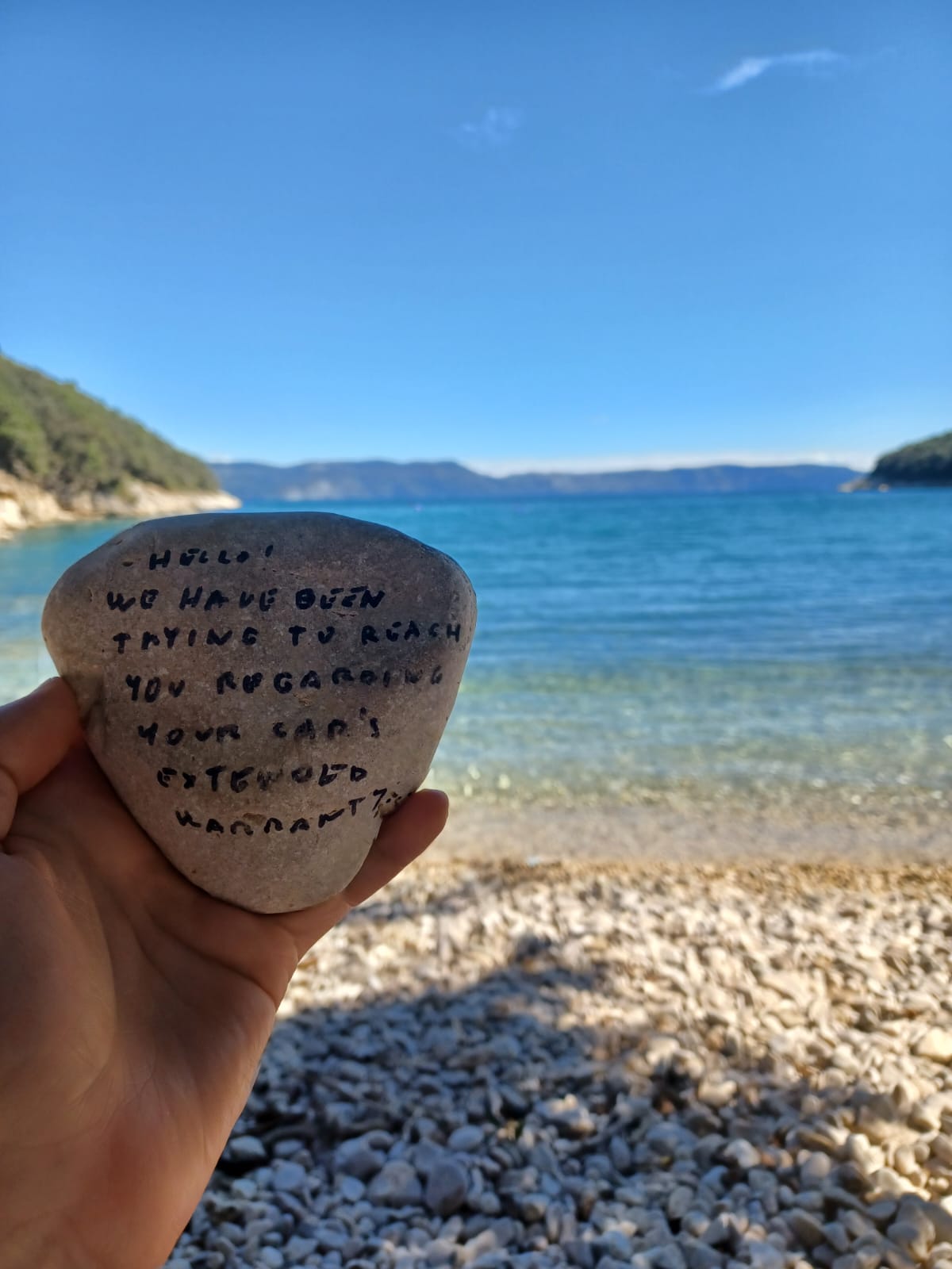 stone in hand, with a beach background.
Stone has written on it: 
Hello, we have been trying to reach you regarding your car's extendet warenty