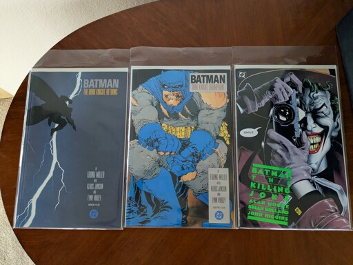 Three comic books sitting on a wooden table: The Dark Knight Returns, The Dark Knight Triumphant, and The Killing Joke